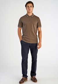 Brun kortärmad polo med dragkedja i kragen och vita detaljer, ihop med marinblå byxor och bruna skor. Slät textur, avslappnad passform.