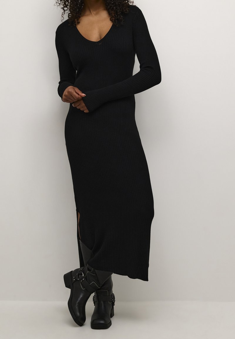 Femme portant une longue robe noire côtelée avec une fente sur le côté et des bottines noires à boucle à la cheville, debout devant un fond clair uni.