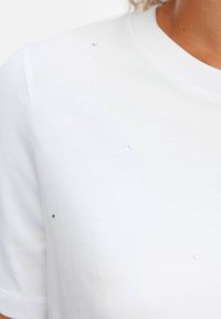 T-shirt bianca a maniche corte con scollatura rotonda, caratterizzata da piccole decorazioni scintillanti sulla parte anteriore per un tocco di dettaglio.