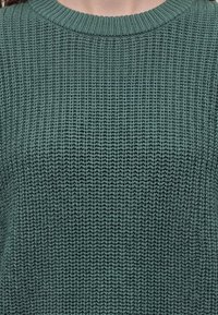 Gros plan d'un pull vert en tricot avec un motif côtelé texturé et un col rond.
