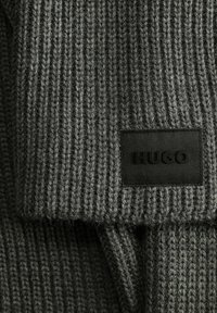 Tejido de punto gris oscuro con acanalado vertical y un parche rectangular negro cosido que muestra el nombre de la marca "HUGO" en letras en relieve.