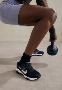 Osoba v černobílých teniskách Nike a šortkách drží černou kettlebelku, zatímco dřepí na světlé podlaze.