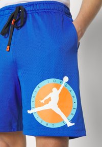 Pantaloni corti in rete blu con elastico in vita, dotati di un laccio nero e un grande logo Air Jordan bianco e arancione sul lato.