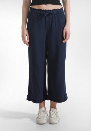 Marineblaue Hosen mit weitem Bein, einem Tunnelzug an der Taille und Rüschenbesatz. Leichter Stoff. Kombiniert mit weißen Sportschuhen.