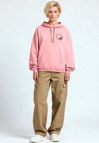 Felpa rosa con una grafica di un cuore e una mano, tasca frontale e polsini elasticizzati. Abbinata a pantaloni cargo beige e scarpe da ginnastica bianche.