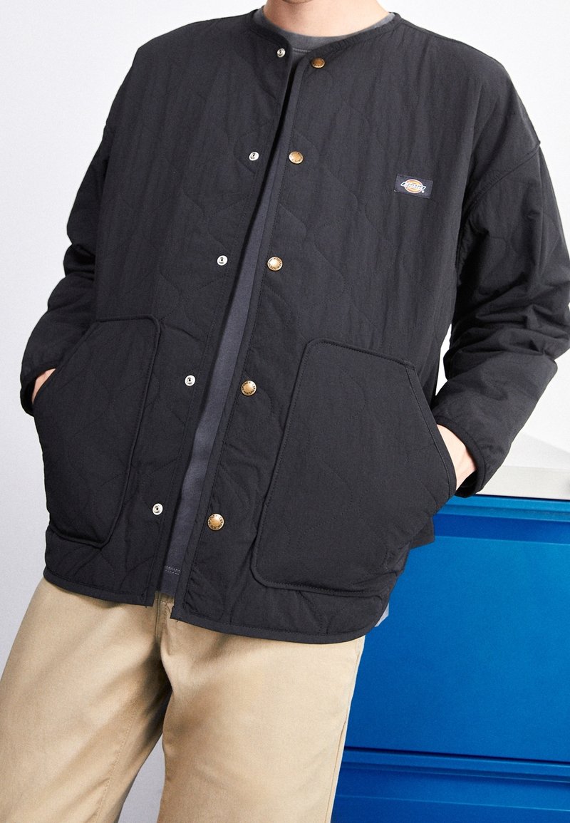 Personne portant une veste matelassée noire avec des boutons-pression et des poches latérales, associée à un pantalon beige, debout à côté d'un meuble bleu.