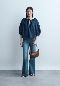 Žena oblečená v tmavomodrej blúzke s nafúknutými rukávmi a modrých džínsoch, drží hnedú kabelku a stojí pred jednoduchým svetlým pozadím.