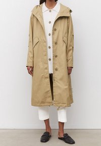 Impermeable beige con capucha, botones a presión, dos bolsillos laterales y un corte holgado. Presenta un cordón en el dobladillo para ajustabilidad.