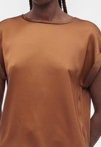 Femme portant un chemisier en satin marron brillant à manches courtes, coupe ample, poignets retroussés et encolure ronde, sur fond uni.