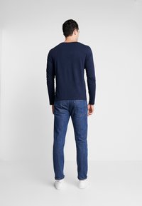 Navyblå långärmad skjorta med rundad halsringning, matchad med åtsittande blå jeans och vita sneakers, sedd bakifrån. Enkel design, utan mönster.