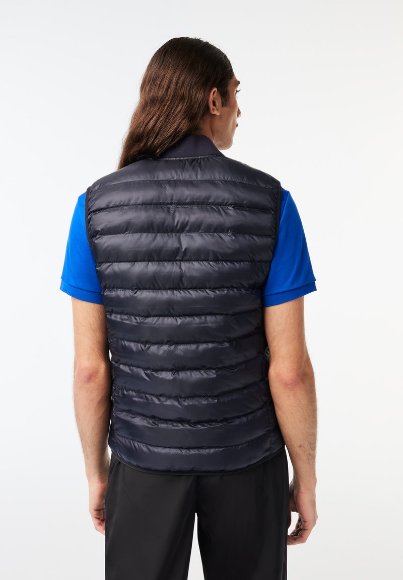 Gilet rembourré navy avec un rembourrage horizontal, un col et des poches latérales, porté sur une chemise bleue, mettant en valeur une texture lisse et brillante.