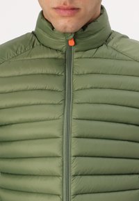 Πράσινο padded μπουφάν με ψηλό γιακά, μπροστινό φερμουάρ και πορτοκαλί λαβή φερμουάρ. Διαθέτει οριζόντια πλιέ κελεί τουαλέτας και λείο υλικό.