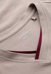 Beige T-shirt med et struktureret stof, med en V-hals og en mørkerød accent på den indre halsudskæring. Etiketten siger "ombre M 14."