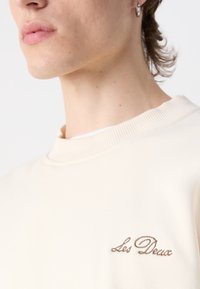 Sudadera color crema con cuello acanalado y texto bordado "Les Deux" en marrón en la parte izquierda del pecho. Textura suave.