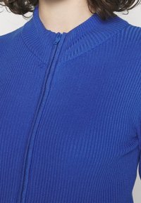 Blå ribbstickad jacka med dragkedja fram och hög krage. Texturen är slät med vertikala linjer skapade av ribbningen.
