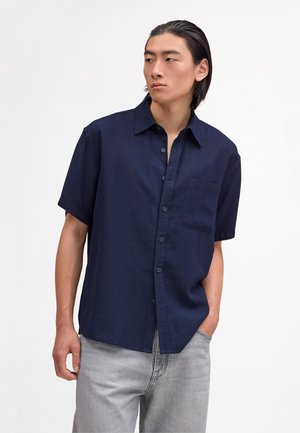 Junger Mann mit glatten dunklen Haaren, der ein marineblaues Hemd mit kurzen Ärmeln und helle graue Jeans trägt, leicht nach links schaut und eine Hand in der Hosentasche hat.