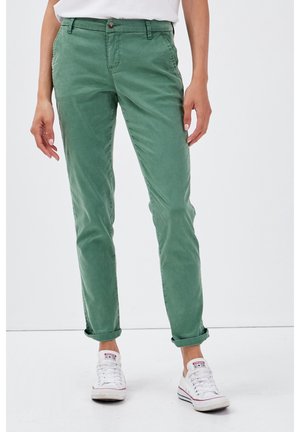 Personne portant un pantalon décontracté vert retroussé et des baskets Converse blanches basses, debout devant un fond clair uni.