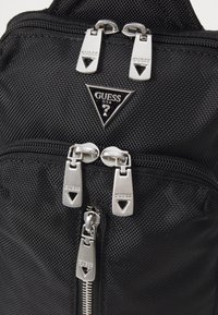 Zwarte textuur nylon rugzak met zilveren hardware ritsen en een driehoekige logo badge. Meerdere met rits afgesloten compartimenten zichtbaar.
