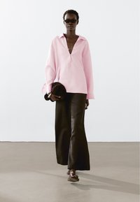 Růžová, oversized blůza s výstřihem do V a dlouhými rukávy, doplněná černými kalhotami s vysokým pasem a širokými nohavicemi. Doplněno texturovanou kabelkou.