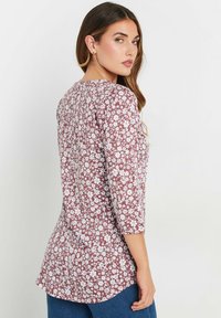 Bordeauxfarbige Bluse mit weißen Blumenmustern, Dreiviertelärmel, Rundhalsausschnitt und abgerundetem Saum. Hergestellt aus leichtem Stoff.
