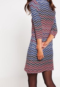 Barevné šaty se zigzag vzorem s tříčtvrtečními rukávy a přiléhavým střihem, vyrobené z texturované, pružné látky v syté červené, modré a bílé barvě.