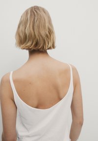 En blond person med kort hår, iført en hvid ærmeløs top, vender væk mod en ensfarvet lys grå baggrund.