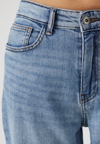 Světle modré džíny z denimu s detailním pohledem, které obsahují přední kapsu, viditelné stehy a kovové zapínání na knoflík v pase.