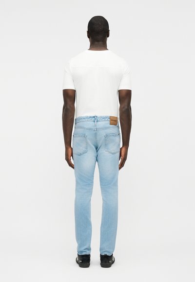 Homme portant une chemise blanche à manches courtes, un jean bleu clair et des chaussures noires, debout de dos devant un fond blanc uni.