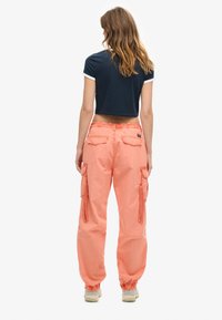 Vrouw met haar rug naar de camera, gekleed in een marineblauwe cropped top, koraalkleurige cargobroek met enkelmanchetten en beige sneakers, op een witte achtergrond.