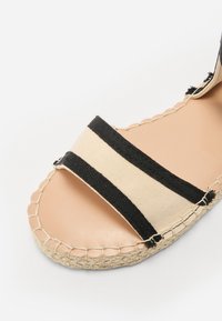 Sandale espadrile cu un superior din pânză bej cu dungi negre, talpă din juta naturală și detalii cusute pe margine.
