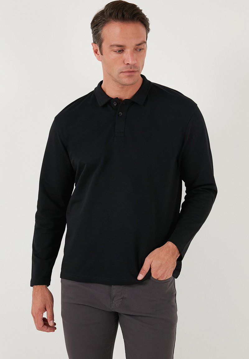 Camisa polo negra de manga larga con cuello, tapeta de tres botones y tela suave. Llevada con pantalones grises, ajustados al torso.