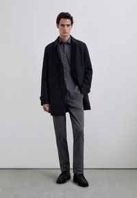 Donkere wollen jas met dubbelgeknoopt ontwerp, gecombineerd met een lichtgrijs overhemd met knopen, grijze broek en zwarte schoenen. Soepele texturen, minimalistische stijl.