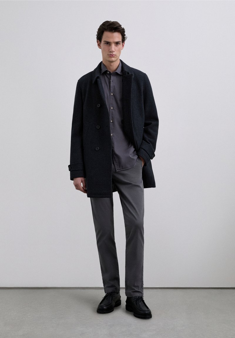 Donkere wollen jas met dubbelgeknoopt ontwerp, gecombineerd met een lichtgrijs overhemd met knopen, grijze broek en zwarte schoenen. Soepele texturen, minimalistische stijl.
