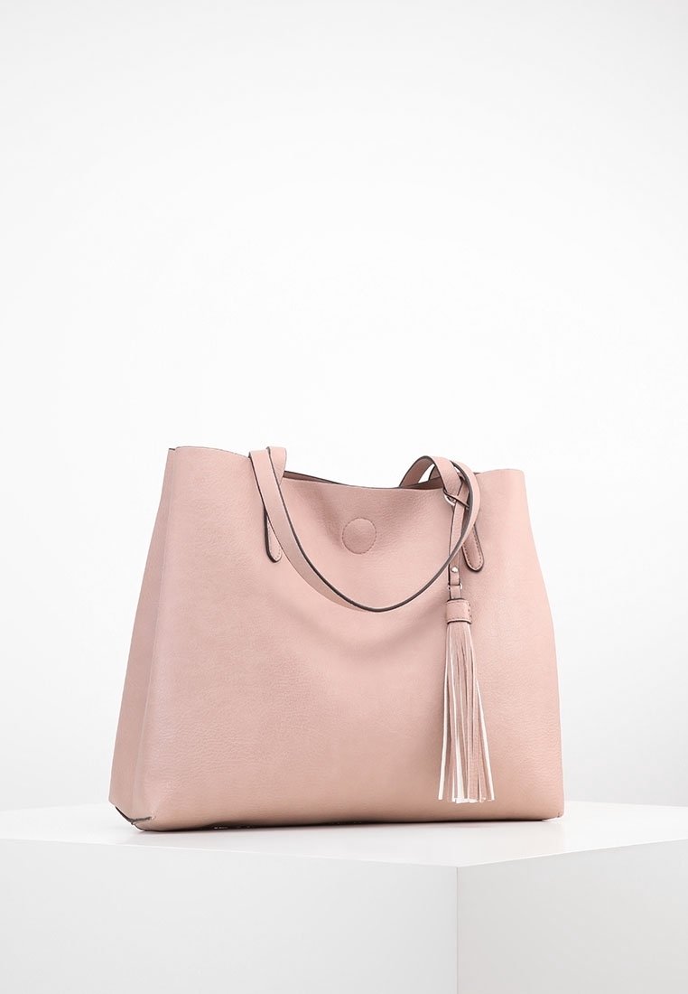 Sac cabas en cuir rose doux avec double poignée et un long détail en pampille, posé sur un socle blanc contre un fond uni.