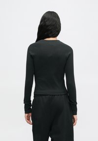 Femme avec de longs cheveux noirs portant un haut noir ajusté à manches longues en côte et un pantalon noir ample, se tenant de dos sur un fond uni.