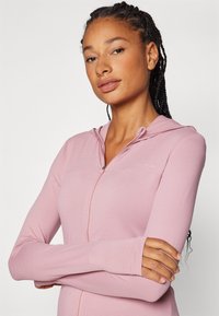 Ljusrosa zip-up jacka med texturerad tyg, figurnära design, huva och långa ärmar. Har tumhål vid ärmsluten.