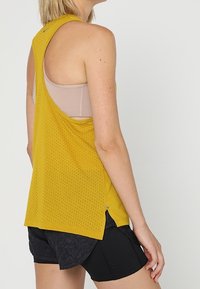 Top giallo perforato con una vestibilità ampia e schiena scoperta, indossato sopra un reggiseno sportivo beige, abbinato a pantaloni corti neri.