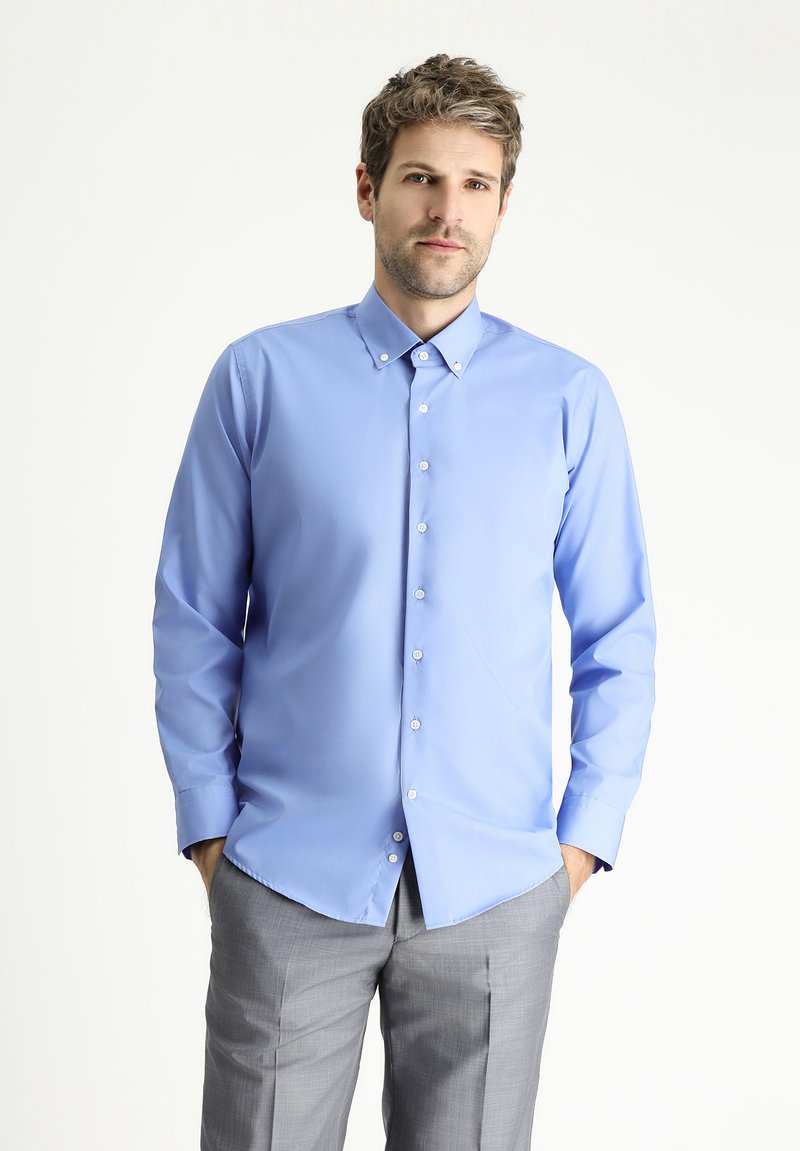 Camicia a maniche lunghe azzurro chiaro con colletto a bottoni e bottoni bianchi, abbinata a pantaloni grigi chiari. Tessuto liscio, vestibilità sartoriale.