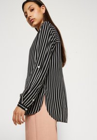 Black and white striped long-sleeve shirt featuring a rounded hem, button-cuff details, and side slits for enhanced mobility.
