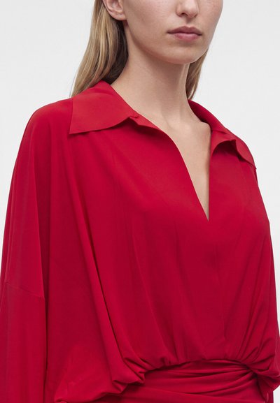 Γυναίκα με φαρδιά κόκκινη μπλούζα με βαθύ V λαιμόκοψη και φαρδιά γιακά σε απλό ανοιχτόχρωμο φόντο.