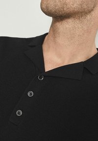 Černé polo tričko z texturované pleteniny s límcem, obsahující tři černé knoflíky a vypasovaný střih. Jednoduchá tkanina bez vzorů.