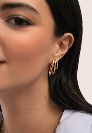 Mujer con cabello oscuro y liso que lleva tres pendientes de aro dorados enlazados y un delicado collar de cadena dorada, sobre un fondo claro.