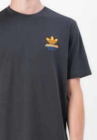 Sötétszürke pamut póló rövid ujjakkal, sárga Adidas logóval és kék szöveggel a bal felső oldalon.