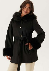 Cappotto nero con collo e polsini in pelliccia sintetica, vita evidenziata da una cintura e chiusura con bottone in argento. Il tessuto ha una texture liscia e una silhouette strutturata.