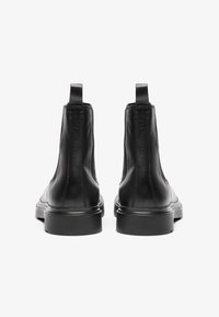 Botas de tobillo de cuero negro con una textura suave, punta redondeada y suela plana. Cuentan con lengüetas traseras y detalles de costura mínimos.