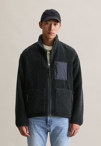 Jeune homme portant une veste polaire noire, une chemise à superpositions vert olive, une casquette noire et un jean bleu clair devant un fond uni.