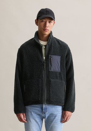Jonge man in een zwarte fleecejack, olijfgroene laagshirt, zwarte pet en lichtblauwe jeans tegen een effen achtergrond.