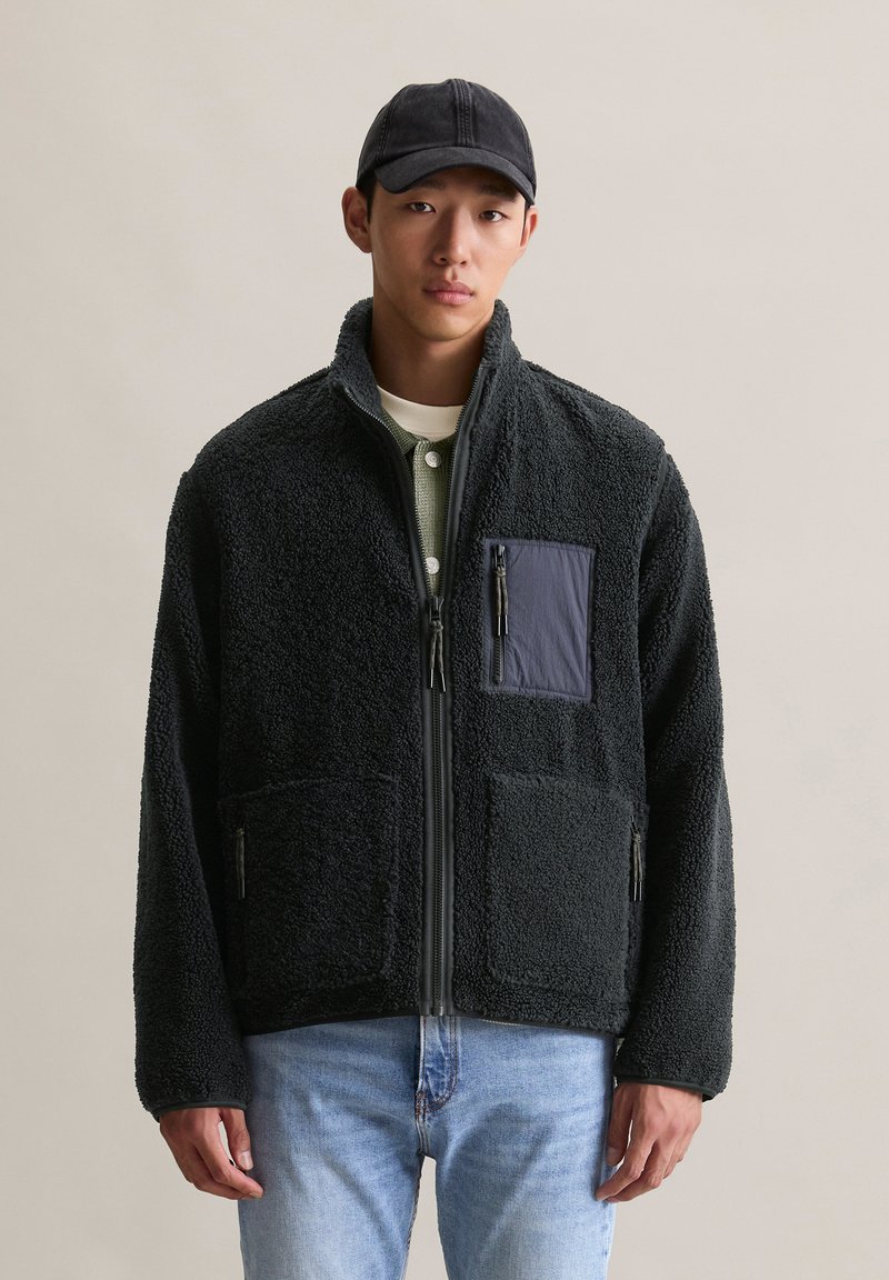 Jeune homme portant une veste polaire noire, une chemise à superpositions vert olive, une casquette noire et un jean bleu clair devant un fond uni.