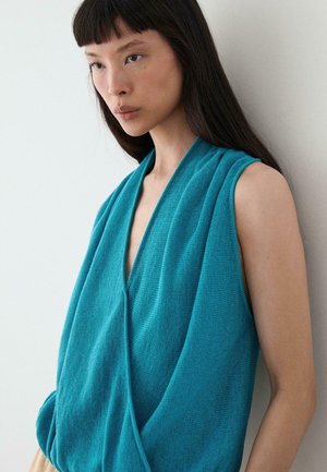 Młoda kobieta z długimi ciemnymi włosami, ubrana w turkusowy, bezrękawnik z drapowaniem, stojąca na tle jasnej, gładkiej ściany, lekko zwrócona w lewo.