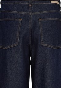 Jean en denim bleu foncé présentant un design classique à cinq poches, avec des coutures visibles et une texture lisse. Comprend une étiquette en cuir à la taille.